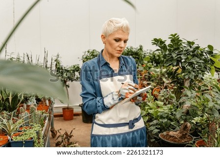 People and greenhouse concept. A woman takes care of the garden. Garden center worker taking notes. Serious woman gardener checking plants holding notebook taking notes about greenery.