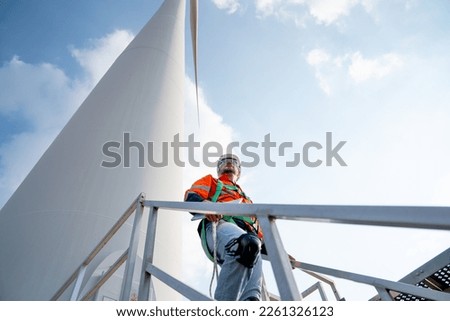 Caucasian engineer or technician walk down at base of windmill or wind turbine and look to right side and blue sky.