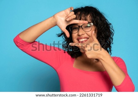 Young exotic brunette woman making a hand frame, composing picture ideas in pink blouse  on a blue studio background. Front view.