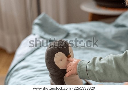 Baby's hand shows and touches the head of a toy penguin, background with space for text.	