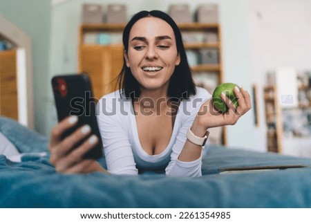 Positive young female smiling and looking at screen of mobile phone while reading text message and relaxing on bed in cozy room at home