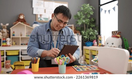 Young chinese man preschool teacher using touchpad sitting on table at kindergarten Royalty-Free Stock Photo #2261369819