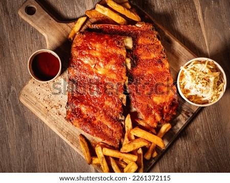 grilled pork ribs with french fries and sauces served on a wooden cutting board, view from top