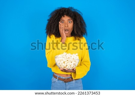 Shocked young woman with afro hairstyle wearing yellow sweater against blue background looks with great surprisment being very stunned, astonished with unexpected news, Facial expressions concept.