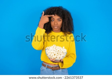 young woman with afro hairstyle wearing yellow sweater against blue background pointing unhappy at pimple on forehead, blackhead  infection. Skincare concept.