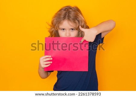 Kid pointing on empty sheet of paper, isolated on yellow background. Portrait of a kid holding a blank placard, poster. Copy space.