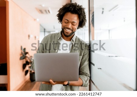 Curly young african-american businessman standing in office hallway and using laptop for office work, male employee checks mail, conducts business correspondence with customers Royalty-Free Stock Photo #2261412747