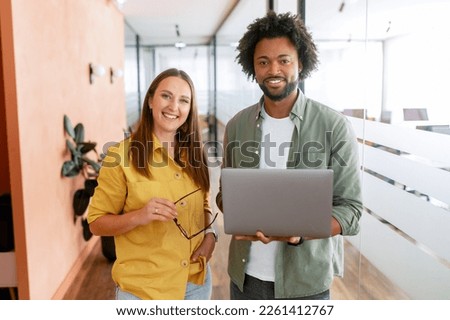Two happy diverse colleagues African American man and Caucasian woman looking at camera standing in office lobby hall, the man is holding laptop Royalty-Free Stock Photo #2261412767