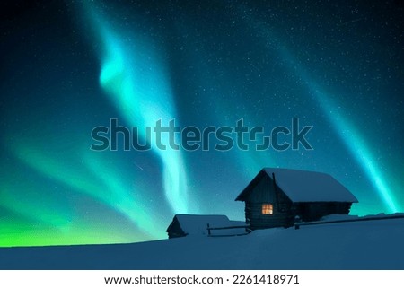 Fantastic winter landscape with wooden house with light in window in snowy mountains and northen light in night sky. Christmas holiday and winter vacations concept Royalty-Free Stock Photo #2261418971