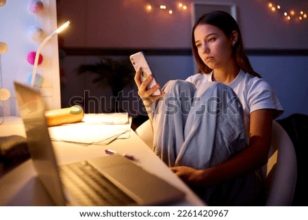 Worried Teenage Girl Sitting At Desk In Bedroom At Home Looking At Mobile Phone At Night Royalty-Free Stock Photo #2261420967