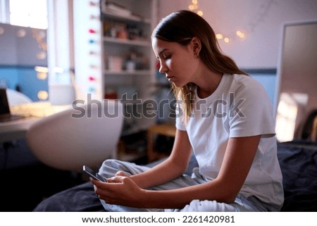 Depressed Teenage Girl Sitting On Bed At Home Looking At Mobile Phone Royalty-Free Stock Photo #2261420981