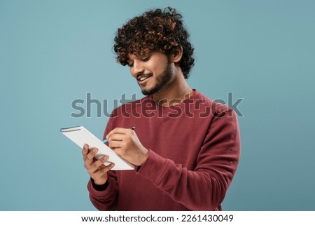 Portrait of smiling Indian man taking notes in notepad isolated on blue background. Smart asian university student studying, learning language, exam preparation. Education concept Royalty-Free Stock Photo #2261430049