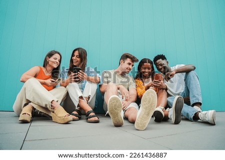 Multiracial group of young friends enjoying and smiling using their mobile phone app sitting at teal wall. Diverse teenagers having fun sharing messages with each other on cellphone at blue clor Royalty-Free Stock Photo #2261436887