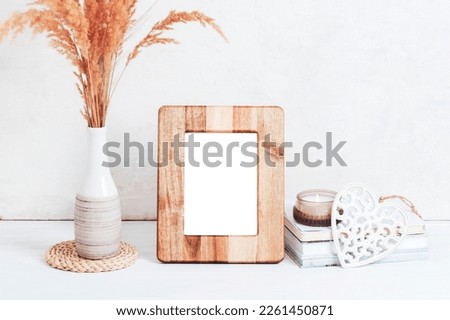 Mockup wooden picture frame, books, aroma candle, carved decor heart and pampas grass in vase on white table. Copy space