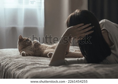 A sad young woman with seasonal affective disorder lies alone on the bed and looks out the window, next to a domestic cat. Concept of winter depression due to lack of sunlight, selective focus Royalty-Free Stock Photo #2261454255