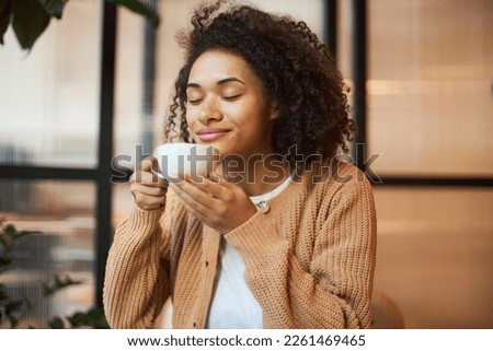 Beautiful smiling African American woman with her eyes closed, enjoying her morning coffee in modern cafe. Coffee break concept Royalty-Free Stock Photo #2261469465