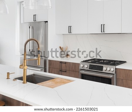 A kitchen detail with a gold faucet, white and wood cabinets, stainless steel appliances, and marble countertops. Royalty-Free Stock Photo #2261503021