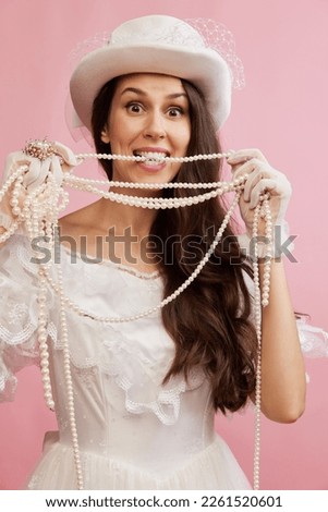 Happy. Portrait of beautiful lady in white vintage dress posing with luxurious pearl necklace over pink background. Concept of 19th century, fashion, comparison of eras, history, retro style