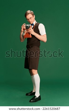 Taking photo with vintage camera. Portrait of man in stylish classical clothes, shorts and vest posing on green studio background. Concept of emotions, facial expression, lifestyle, retro fashion. Ad