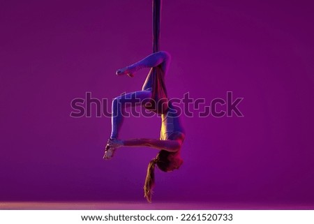 Hanging upside down. Young flexible girl doing aerial yoga, training on purple studio background in neon light. Concept of fitness, sportive lifestyle, health, strength, aerial yoga, anti-gravity yoga