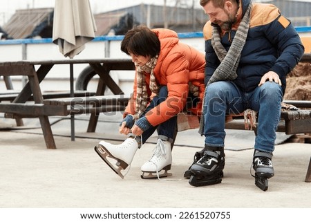 Mature man and woman getting ready to skate on ice rink outdoors. Woman tying laces on white skates. Active weekends. Concept of leisure activity, winter hobby, vacation, fun, relationship, emotions.