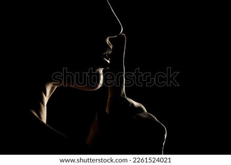 Silhouette of unknown woman with the face in the shadow holding finger on her lips on a black studio background Royalty-Free Stock Photo #2261524021