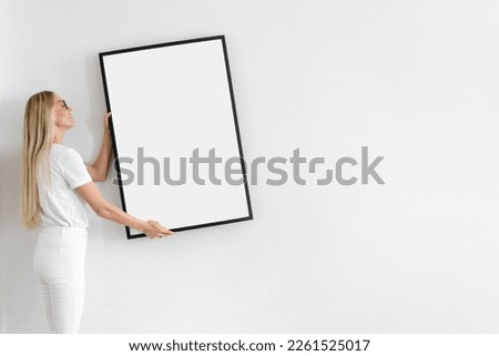 smiling female holding blank poster in frame and decorate wall on white background in living room, domestic interior concept Royalty-Free Stock Photo #2261525017