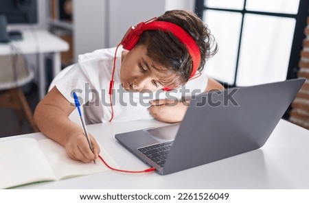 Adorable hispanic boy student using laptop writing on notebook at classroom