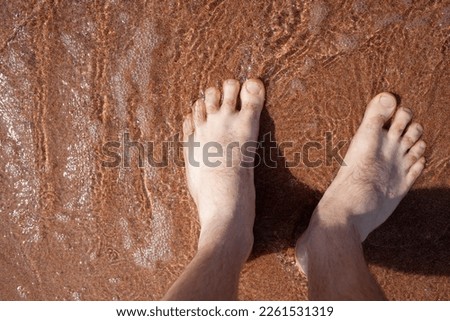 Top view of men's legs in the sea. A man is standing on the sand. An ocean wave washes the shore. Summer holidays.