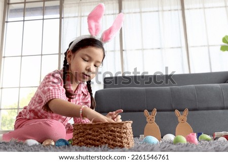 Happy adorable pink little Easter bunny girl kid with rabbit ears headband preparing beautiful colorful painted Easter egg basket in living room, decorations to celebrate of Easter spring beginning.
