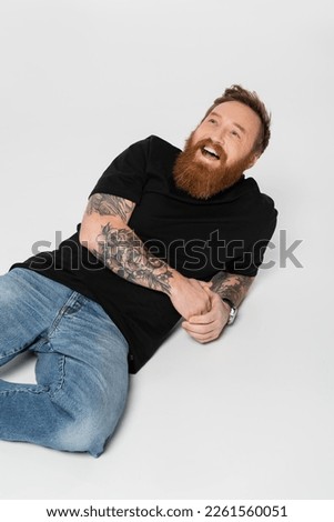 joyful bearded and tattooed man in jeans and black t-shirt laughing while lying on grey background
