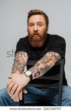 thoughtful bearded man in black t-shirt and wristwatch sitting on chair and looking at camera isolated on grey