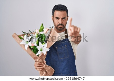 Hispanic man with beard working as florist pointing with finger up and angry expression, showing no gesture 