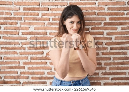 Young brunette woman standing over bricks wall ready to fight with fist defense gesture, angry and upset face, afraid of problem 