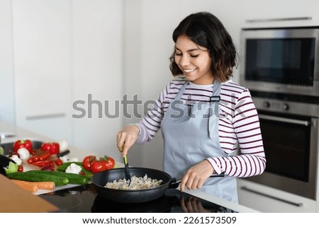 Beautiful young middle eastern woman holding cooking spatula, mixing rice with vegetables in frying pan on electric stove, preparing food at kitchen, copy space. Healthy diet, nutrition