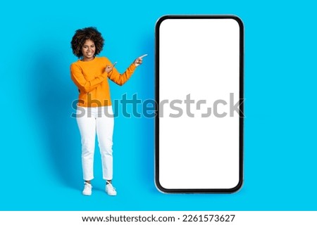 Happy pretty stylish young black woman with nice hairstyle in casual pointing at huge cell phone with white blank screen, mockup for online offer, isolated on blue studio background, copy space