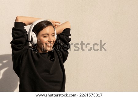 Cheerful caucasian millennial female in sportswear, wireless headphones do stretching exercises for arms and look at empty space on white wall background. Sports and music, body care and weight loss