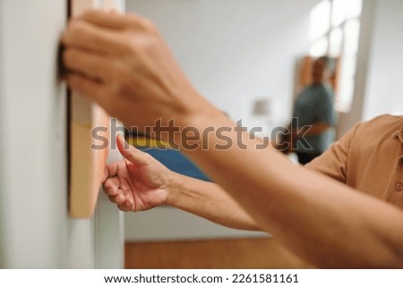 Hands of aged man hanging picture on wall to decorate apartment