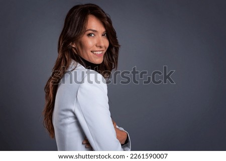 Close-up of an attractive middle aged woman with toothy smile wearing white blazer while sitting at isolated dark background. Copy space. Studio shot.