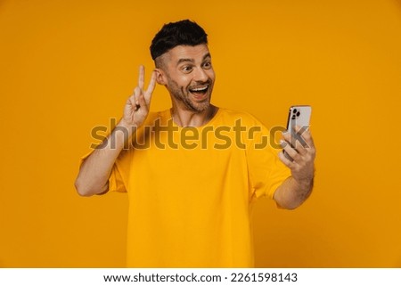 Cheerful bearded man in yellow t-shirt making video call via mobile phone and showing peace sign isolated over yellow studio wall