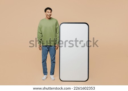 Full body surprised young man of African American ethnicity wear green sweatshirt look at big huge blank screen mobile cell phone smartphone with area isolated on plain pastel light beige background