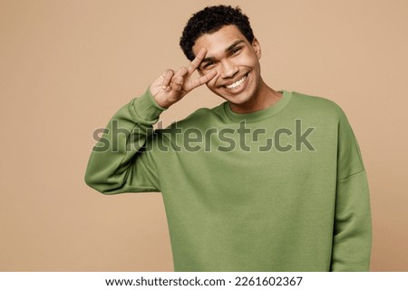 Young smiling fun man of African American ethnicity wear green sweatshirt show cover cover showing victory sign isolated on plain pastel light beige background studio portrait People lifestyle concept