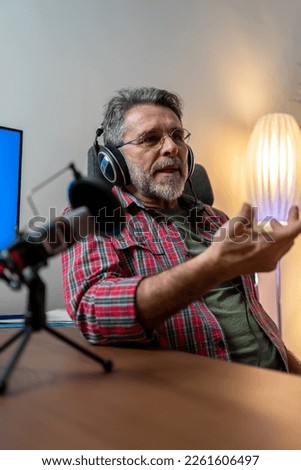 A mature man host streaming his audio podcast using microphone and laptop at his small broadcast studio, vertical. Smiling radio host in headphones recording podcast in broadcasting studio