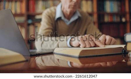 Senior historian reading a book and typing notes on laptop, working on an article Royalty-Free Stock Photo #2261612047