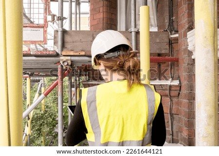Female chartered civil engineer working in a construction site building, taking notes on her tablet, condition inspection survey, hard hat and yellow high visibility vest, photo from behind