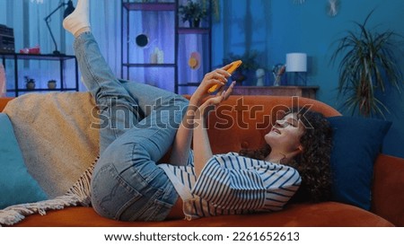 Young woman with curly hairstyle lying on sofa uses mobile phone smiles at night evening home. Girl texting share messages content on smartphone social media applications online watching relax movie Royalty-Free Stock Photo #2261652613