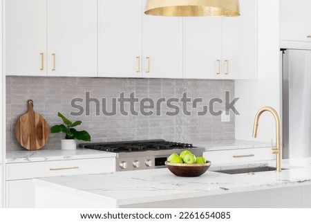 A kitchen detail with white cabinets, gold faucet and light hanging over the island, and a tiled backsplash. Royalty-Free Stock Photo #2261654085