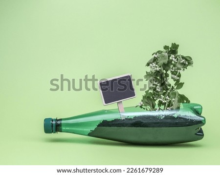 A plastic bottle filled with earth into which a branch with wilted leaves is stuck and a small blank signpost with a place for text lie on a green background, close-up side view.