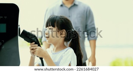Focus progressive young girl holding EV charger device from home electric charging station for renewable energy powered car with blur asian man background. Sustainable green energy technology concept.