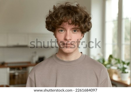 Close up of a young handsome positive curly man with blue eyes with a smiling face wearing casual beige t-shirt looking at camera and standing alone in morning in the modern kitchen at home. Royalty-Free Stock Photo #2261684097